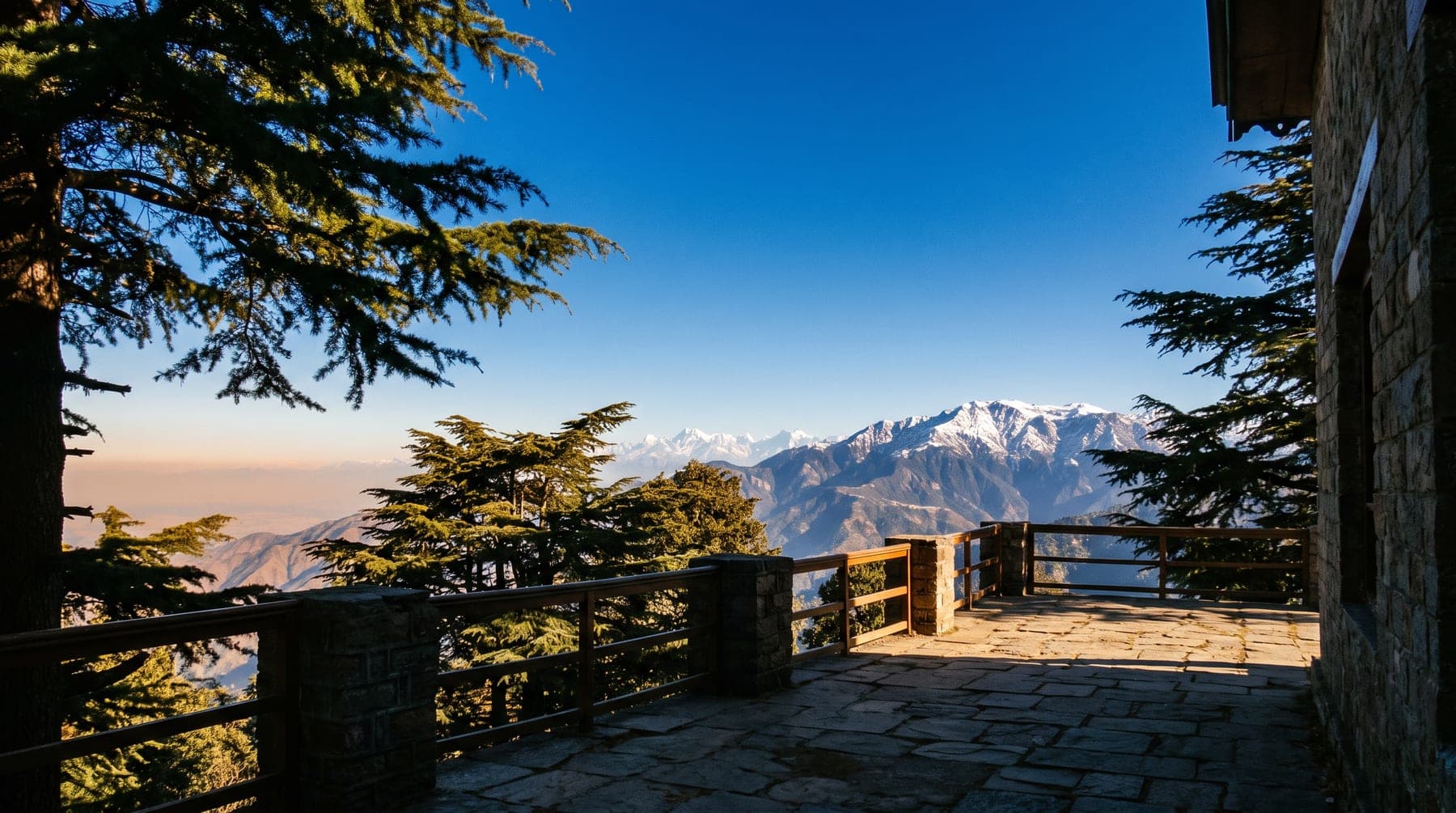 A stone terrace under deodar looking out to a snow-covered Himalayan range on a clear morning